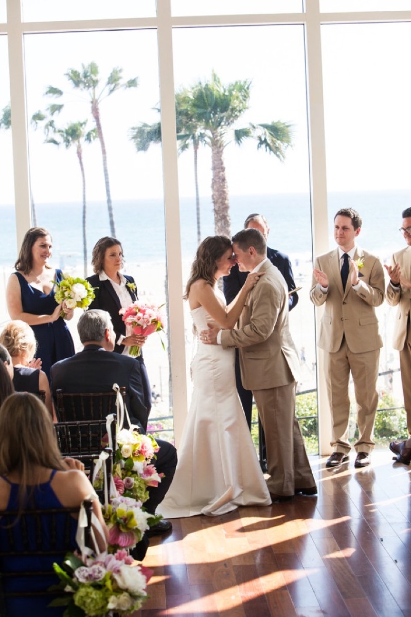 Bride and groom touching foreheads during the ceremony with wedding party and beach in the window