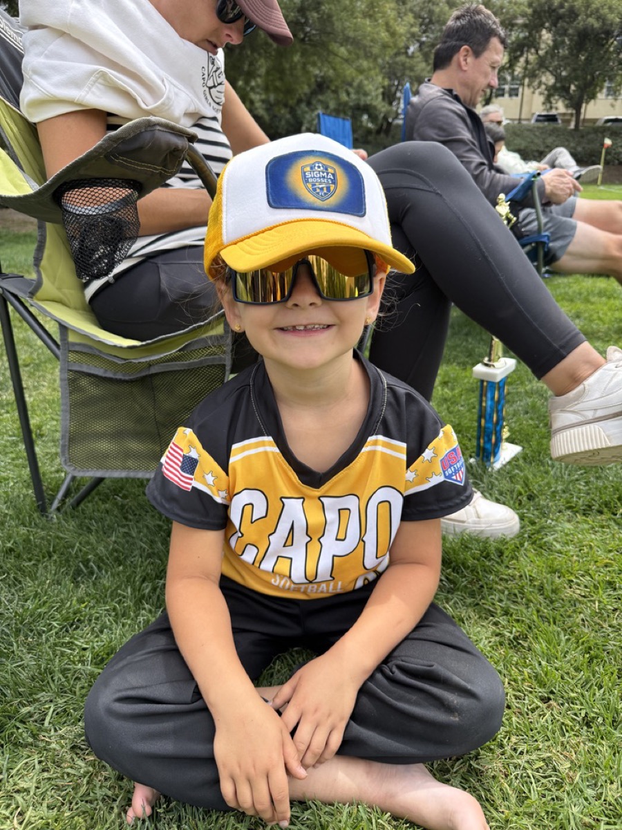 Morgan in softball gear with an oversized hat and mirrored sunglasses on the grass